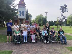Pokaż zdjęcie: na zdjęciu grupa osób w parku w oddali drzewa i plac zabaw, osoby pozują do zdjęcia na pierwszym planie osoby na wózku inwalidzkim w drugim rzędzie osoby stojące 