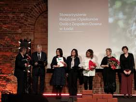 Pokaż zdjęcie: Na scenie stoją elegancko ubrani ludzie. Jeden z panów przemawia do mikrofonu. Niektóre kobiety trzymają w dłoniach kwiaty. W tle widać napis: Stowarzyszenie Rodziców i Opiekunów Osób z Zespołem Downa.