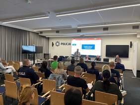 Pokaż zdjęcie: Na Sali konferencyjnej na drewnianych krzesłach siedzi wiele ludzi. Przed nimi szeroki stół za którym stoi mężczyzna ubrany w jasnoszarą marynarkę. Przy stole siedzi jeszcze dwóch mężczyzn (jeden w stroju służbowym policjanta) i kobieta. Za n