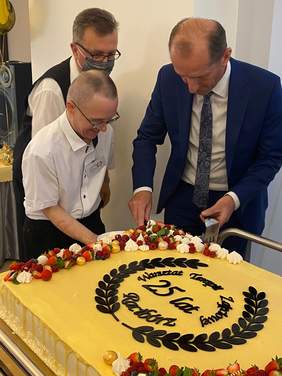 Pokaż zdjęcie: Na pierwszym planie widać duży tort w kształcie prostokąta na żółtym tle ciemnobrązowy napis Warsztat Terapii Zajęciowej w Raciborzu 25 lat. Z boku stoi trzech mężczyzn, jeden z nich kroi tort drugi podaje talerz. 