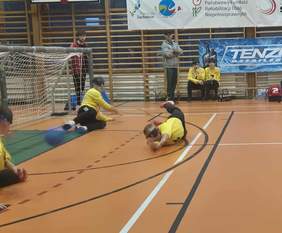 Sala sportowa. Osoby w żółtych koszulkach, prawdopodobnie zawodnicy goalball, leżą lub klęczą na pomarańczowej podłodze w pobliżu bramki. Jedna osoba rzuca się po piłkę. Po prawej stronie widać trenera w czarnej koszulce w dynamicznej pozie. W tle widoczne bannery sponsorów i organizatorów w języku polskim, w tym "Państwowy Fundusz Rehabilitacji Osób Niepełnosprawnych" i "Ministerstwo Sportu i Turystyki".