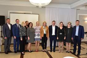 Pokaż zdjęcie: Na scenie przed ekranem, stoi w rzędzie grupa 11 osób elegancko ubranych pozujących do zdjęcia