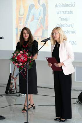 Pokaż zdjęcie: Dwie kobiety stoją przy mikrofonach. Kobieta po lewej trzyma bukiet czerwonych i różowych kwiatów, ubrana w czarną sukienkę, a kobieta po prawej, w jasnej marynarce i czarnych spodniach, przemawia do publiczności, trzymając teczkę. W tle plakat z aniołem.