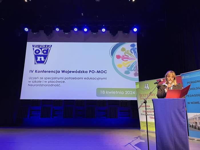 Pokaż zdjęcie: Slajd z prezentacji wyświetlony na pulpicie, widać salę konferencyjną, obok mówinica, opowiada kobieta, 