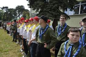 Pokaż zdjęcie: W rzędzie stoją dzieci: druhny i druhowie. Mają na sobie harcerskie ubrania. W tle widać budynek i drzewa. 