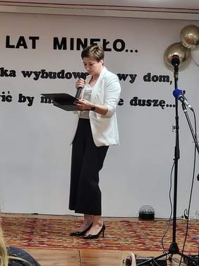 Pokaż zdjęcie: Na scenie stoi młoda kobieta z krótkimi włosami, ubrana w białą marynarkę i czarne spodnie, w ręku trzyma mikrofon oraz drewniany grawerton. 