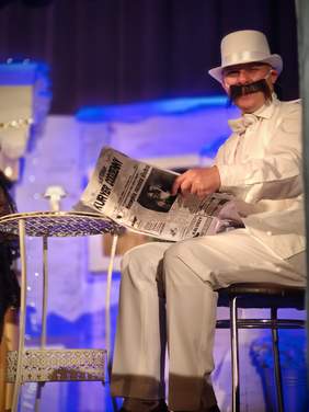 Pokaż zdjęcie: Mężczyzna w białym kostiumie, cylindrze i z dużym sztucznym wąsem siedzi przy stoliku, trzymając gazetę. Scena ma teatralny charakter, z niebieskim oświetleniem w tle.