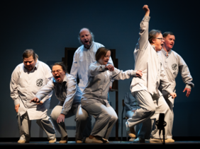 Pokaż zdjęcie: Na scenie stoi kilka osób ubranych w jasne fartuchy z wyszywanym logo na piersi w kształcie litery B. Wszyscy mają prawdopodobnie krzyczą. Każdy patrzy w innę stronę i ma inną pozę.