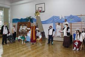 Pokaż zdjęcie: Na scenie stoi mężczyzna z rozłożonymi rękami. Obok niego siedzi kobieta z dzieckiem  na ręku.Za mężczyzną szopka  ze zwierzętami oraz 2 kobiety i mężczyzna  przebrani   za anioły. Obok nich stoi kobieta i mężczyzna przebrany za pastuszka. 