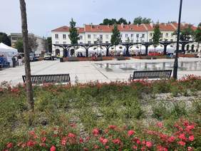 Pokaż zdjęcie: Zdjęcie przedstawia Stary Rynek w Łodzi. Na środku znajduje się plac z fontannami, wokół którego ustawione są ławki dla spacerowiczów. W tle widać stoiska wystawiennicze. 