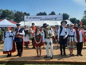 Pokaż zdjęcie: Na podeście terenu rekreacyjnego stoją ludzie ubrani na ludowo. Tworzą pary, trójki lub większe podgrupy. Są ubrani w kolorowe folklorystyczne stroje. 