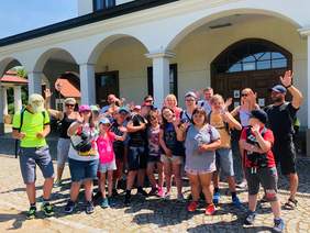 Pokaż zdjęcie: Słoneczny dzień. Grupa uczestników wyjazdu kolonijnego, opiekunowie oraz dyrektor Marta Mordarska stoją na placu rynkowym w Miasteczku Galicyjskim w Nowym Sączu. Wszyscy uśmiechają się i machają. W tle fragment zabytkowego budynku