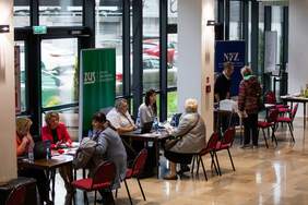 Pokaż zdjęcie: Fragment pomieszczenia z przeszkloną ścianą pod którą ustawione są trzy stoliki, siedzą przy nich kobiety i rozmawiają