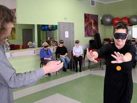 Pokaż zdjęcie: Ubrana na czarno kobieta w spiętych ciemnych włosach ma opaskę na oczach. Idzie z wyciągniętymi rękoma. Na przeciw mężczyzna wyciągający do niej ręce. Do dokoła siedzą osoby z opaskami na oczach.