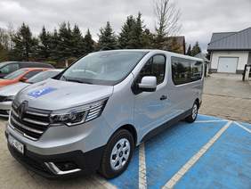 Pokaż zdjęcie: Zdjęcie przedstawia srebrny bus marki Renault, zaparkowany na miejscu dla osób z niepełnosprawnościami, oznaczonym niebieską nawierzchnią i symbolem wózka inwalidzkiego. Na masce pojazdu widnieje również niebieska naklejka z tym samym symbolem. 