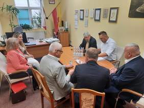 Pokaż zdjęcie: Przy owalnym stole siedzi siedem osób: dwie kobiety i pięciu mężczyzn. Podpisują umowę. Na stole leżą różne dokumenty. W tle na ścianie wiszą różne dyplomy. 