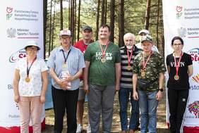 Pokaż zdjęcie: Grupa uczestników Wojewódzkiej Spartakiady Osób Niepełnosprawnych 2024 w Ogrodniczkach pozuje do zdjęcia z medalami na tle banerów sponsorów i organizatorów.