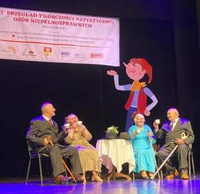 Pokaż zdjęcie: Na scenie przy stoliku siedzą dwie starsze kobiety i dwóch starszych panów. W prawych dłoniach trzymają filiżanki a w lewych laski. Za nimi postać Pinokia, a nad nimi wisi baner z napisem Przegląd Twórczości Artystycznej Osób Niepełnosprawnych