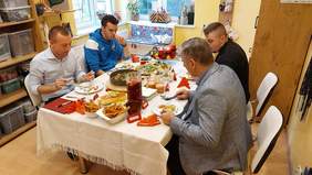 Pokaż zdjęcie: Przy stole siedzi czterech mężczyzn konsumujących potrawy. Na stole leży kilka potraw, napój w dzbanku, patera z pieczoną potrawą i deserki w słoikach.