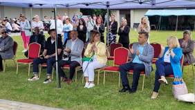 Pokaż zdjęcie: Na trawie, stoją czerwone krzesła, siedzą na nich elegancko ubrani ludzie i klaszczą. Nad nimi dach namiotu. 