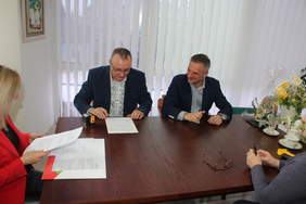 Pokaż zdjęcie: W gabinecie przy ciemnym okrągłym stole siedzą 4 osoby, dwóch mężczyzn i dwie kobiety. Mężczyźni maja przed sobą dokumenty, jeden z nich podpisuje umowę. Kobieta siedząca po lewej stronie stołu, blondynka w czerwonej marynarce układa dokumenty w teczce.