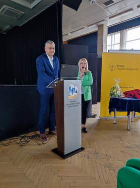 Pokaż zdjęcie: Podczas konferencji przy mównicy stoi mężczyzna i kobieta, obecnie przemawia kobieta w zielonej marynarce.