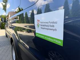 Zbliżenie na tabliczkę umieszczoną na granatowym mikrobusie. Widoczne są logo i pełna nazwa Państwowego Funduszu Rehabilitacji Osób Niepełnosprawnych oraz napis na zielonym pasku: „Dofinansowane ze środków PFRON”.