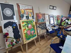 Przestronna sala. Wzdłuż ścian rozmieszczono wiele obrazów na sztalugach, krzesłach. W centralnej części na stołach rozłożone rzeźby i płaskorzeźby.