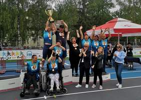 Pokaż zdjęcie: Na podium sportowym stoi zwycięska drużyna. Zawodnicy trzymają w rękach puchar, który podnoszą. Sportowcy są uśmiechnięci i wyciągają ręce do góry. 