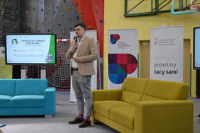 Pokaż zdjęcie: Mężczyzna trzymając mikrofon wygłasza przemówienie na tle grafik promujących PFRON i ZPE w Olsztynie. Po lewej stronie na ekranie telewizora wyświetlana jest prezentacja