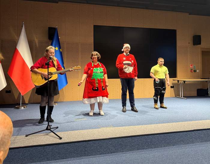 Pokaż zdjęcie: Scena z występu artystycznego. Cztery osoby stoją na podwyższonej scenie na tle trzech flag: po lewej flaga z czerwonym gryfem na białym tle (herb województwa zachodniopomorskiego), obok flaga Polski (biało-czerwona), a po prawej flaga Unii Europejskiej (niebieska z żółtymi gwiazdami). Po lewej stronie sceny młoda dziewczyna gra na gitarze i śpiewa do mikrofonu. Ma na sobie czerwoną bluzkę i czarną spódnicę/spodnie, a jej włosy są związane w kucyk. Obok niej stoi kobieta w fartuchu z motywem choinki i białej spódnicy, uśmiechnięta. Po prawej stronie sceny dwóch mężczyzn, jeden w świątecznym swetrze z Mikołajem i czapce Mikołaja, a drugi w żółtej koszulce polo i ciemnych sportowych spodniach. W dolnym lewym rogu widać fragment głowy i ramienia widza, siedzącego na widowni. Kontekst sugeruje wydarzenie świąteczne lub przedświąteczne, prawdopodobnie w budynku administracji publicznej lub sali konferencyjnej.