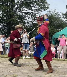 Pokaż zdjęcie: Dwóch mężczyzn ubranych w stroje historyczne walczy szablami na oczach publiczności. Jeden z nich nosi czerwony kontusz i futrzaną czapkę, drugi kolorowy strój z niebieskimi i zielonymi elementami oraz piórem w czapce. W tle widać grupę widzów oraz zielony namiot.