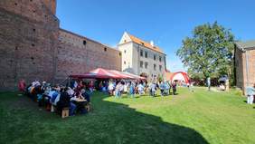 Pokaż zdjęcie: Na dziedzińcu Zamku Królewskiego w Łęczycy znajdują się rozłożone namioty. Pod nimi jest scena i widownia. Wokół stoją ludzie. 