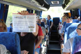 Pokaż zdjęcie: W autokarze po prawej i lewej stronie siedzą pasażerowie. Są to kobiety i mężczyźni w różnym wieku. Jeden pan pokazuje kartkę z informacją,  że wyjazd jest dofinansowany ze środków PFRON. 