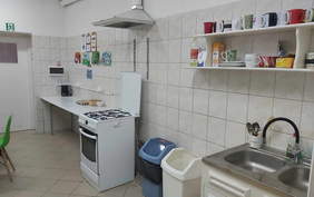 Pokaż zdjęcie: Zdjęcie przedstawia kuchnię przed remontem. Pomieszczenie było z białymi kafelkami na ścianach i jasną podłogą. Po lewej stronie jest długi biały stół, a na nim czajnik elektryczny. Przy blacie stoi biała kuchenka gazowa z piekarnikiem, nad nią metalowy okap. Obok kuchenki widać dwa kosze na śmieci: jeden niebieski i jeden biały. Po prawej stronie znajduje się metalowy zlew dwukomorowy z baterią. Nad zlewem są dwie półki z kubkami, talerzami i różnymi produktami spożywczymi. 