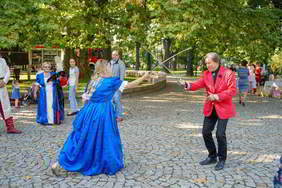 Kobieta o blond włosach w niebieskiej długiej sukni i mężczyzna w czarnych spodniach i czerwonej marynarce walczą na miecze oczywiście na niby, uśmiechają się. Przygląda się tej scenie kilka osób. Wszystko na tle zielonych drzew, podłoże wy