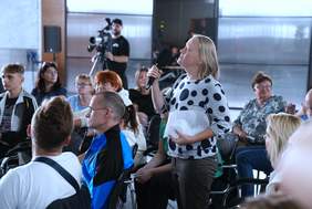 Pokaż zdjęcie: Zdjęcia podczas nagrania, jedna z uczestniczek programu stoi, trzyma w ręku mikrofon i zadaje pytanie gościom. Pozostałe osoby siedzą.