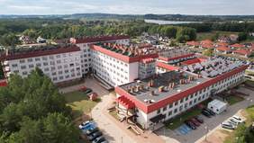 Pokaż zdjęcie: Z lotu ptaka widać wielosegmentowy budynek kilkupiętrowy z podjazdami, drogą i parkingiem dla osób niepełnosprawnych. Na 5 najwyższych dachach z 8 segmentów budynku stoją panele słoneczne