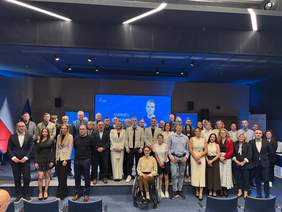 Pokaż zdjęcie: Zdjęcie grupowe wykonane na tej samej scenie, przedstawiające około 30 osób – kobiet i mężczyzn w różnym wieku, ubranych w stroje wizytowe i casualowe. W centralnym punkcie pierwszego rzędu siedzi kobieta na wózku inwalidzkim. W tle na dużym ekranie widoczne jest zdjęcie młodego mężczyzny i napis „DANIEL”. Po lewej stronie sceny stoją flagi Polski oraz Unii Europejskiej. Pomieszczenie jest nowocześnie oświetlone listwami LED na suficie.