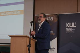 Pokaż zdjęcie: Przy mównicy stoi mężczyzna z siwą brodą, kucykiem i okularach ubrany w ciemny garnitur