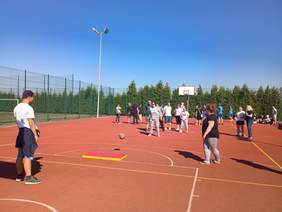 Pokaż zdjęcie: Na tle bezchmurnego nieba widoczna jest grupa kilkunastu osób, która rywalizuje ze sobą na boisku ogrodzonym wysokim płotem. Nisko nad murawą boiska unosi się piłka do gry w siatkówkę.