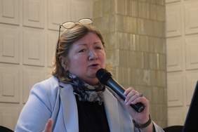 Pokaż zdjęcie: Kobieta z okularami na głowie trzymająca mikrofon w ręku, siedzi za stołem konferencyjnym