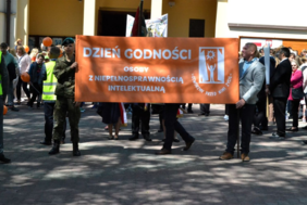 Pokaż zdjęcie: Przed wejściem do kościoła ustawia się grupa osób, na froncie stoją dwaj mężczyźni trzymający pomarańczowy baner z hasłem: Dzień Godności Osoby z Niepełnosprawnością Intelektualną.