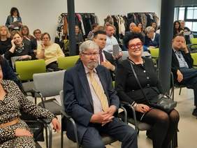 Pokaż zdjęcie: W sali konferencyjnej siedzą na krzesłach uczestnicy konferencji. Niektórzy się uśmiechają, a inni rozmawają przez telefon. 