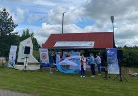 Pokaż zdjęcie: Kilkanaście uśmiechniętych osób ubranych w białe koszulki stoi na tle jeziora. Kilka osób ma na głowie marynarskie czapki