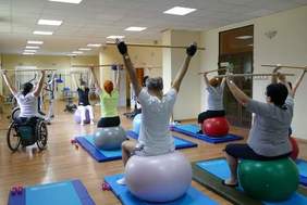 Pokaż zdjęcie: W sali gimnastycznej ćwiczą różne osoby. Jedna pani siedzi na wózku inwalidzkim, a inni na piłkach gimnastycznych. Wszyscy trzymają ręce uniesione pionowo w górę. 