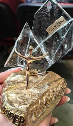 Pokaż zdjęcie: Zbliżenie na statuetkę Lodołamacze – złotą figurkę człowieka z uniesionymi ramionami, stojącą na podstawie z napisem „Lodołamacze”, z elementami przypominającymi kryształowe bryły lodu.