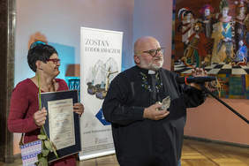 Pokaż zdjęcie: Przed mikrofonem stoi ksiądz w okularach. W ręku trzyma statuetkę. Obok niego kobieta w okularach trzyma certyfikat oprawiony w aluminiową ramę. Za nimi baner z napisem zostań lodołamaczem. 