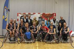 Pokaż zdjęcie: Grupowe zdjęcie drużyny koszykarzy na wózkach Start Białystok. Sportowcy ubrani są w czarne stroje sportowe, siedzą na wózkach, trzymając medale i puchar. Za nimi stoją trenerzy, organizatorzy i goście. W tle widoczne jest logo Lotto, baner z motywem koszykarskim oraz siatka ochronna hali sportowej.