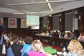 Pokaż zdjęcie: Zdjęcie sali konferencyjnej, stół ustawiony w podkowę, przy nim słuchacze. Prelegent kobieta. W tle slajd z prezentacją. 